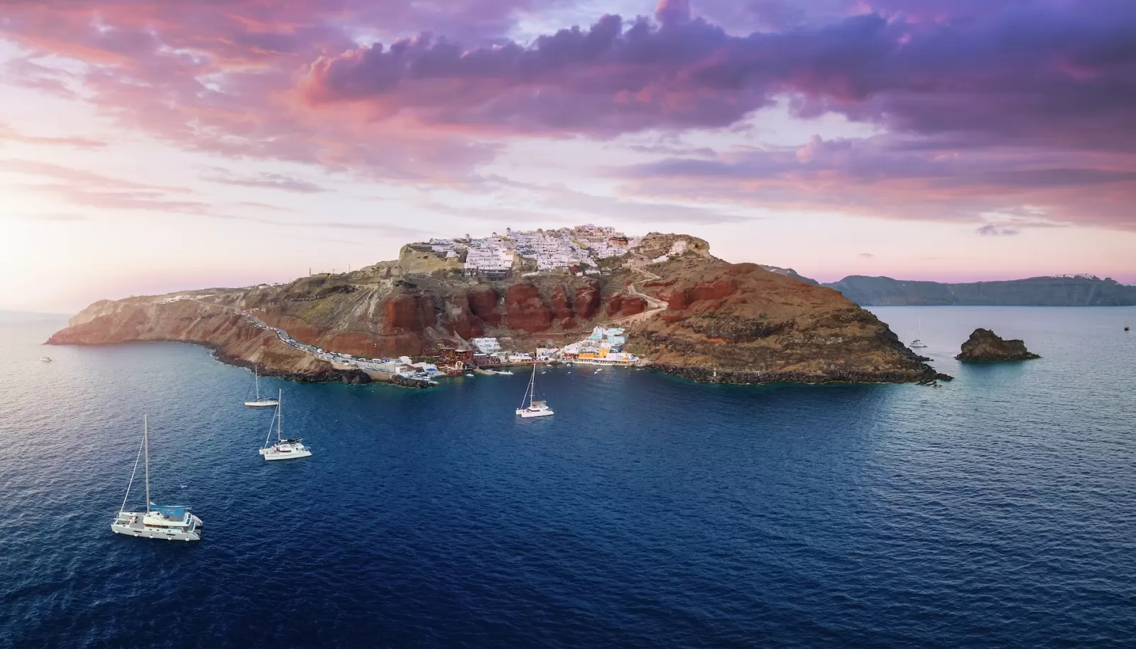Panoramic aerial view to the villages of Oia and Ammoudi surrounded by the steep, volcanic rock formations on Santorini island, Greece, during summer sunset time
